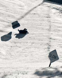 Kid sledding in a blue recycling bin and crashing at the bottom of the hill. There is diamond shaped sign along the bottom of the the path that says "SLOW"