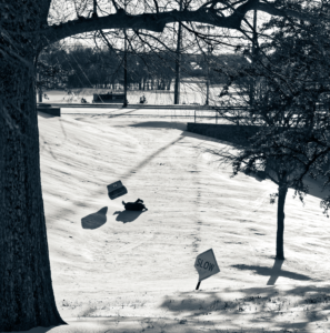 Kid sledding in a blue recycling bin and crashing at the bottom of the hill. There is diamond shaped sign along the bottom of the the path that says "SLOW"