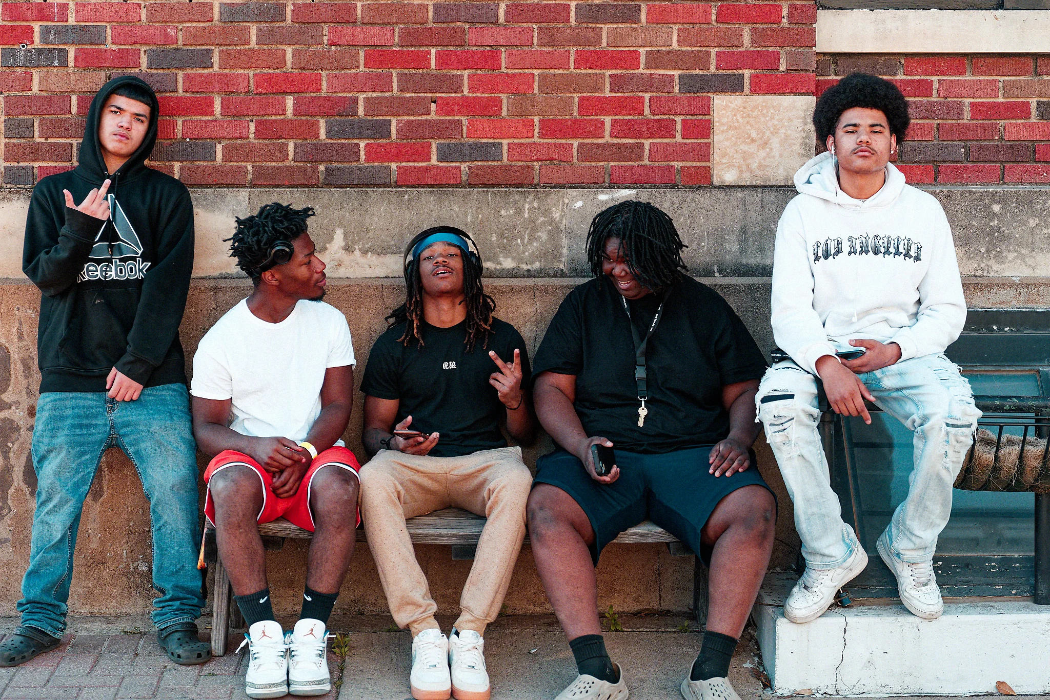 Four young people sit and stand in front of a red brick wall, with one seated on a wooden bench and others leaning or sitting beside it.