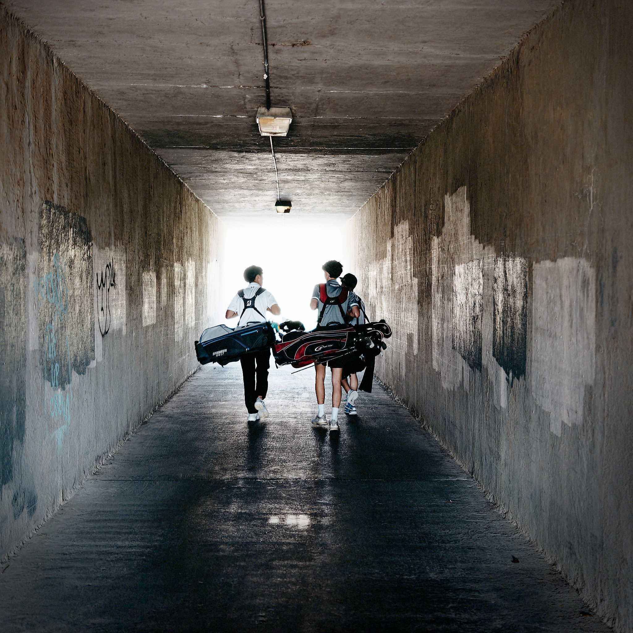 Three young people carry golf bags through a concrete underpass toward a bright opening, with graffiti-marked walls on both sides.