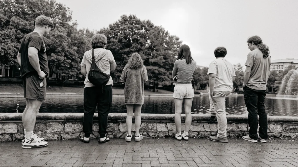 Six people stand in a row with their backs to the camera, looking over a stone wall at a pond with a fountain and trees.