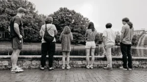 Six people stand in a row with their backs to the camera, looking over a stone wall at a pond with a fountain and trees.