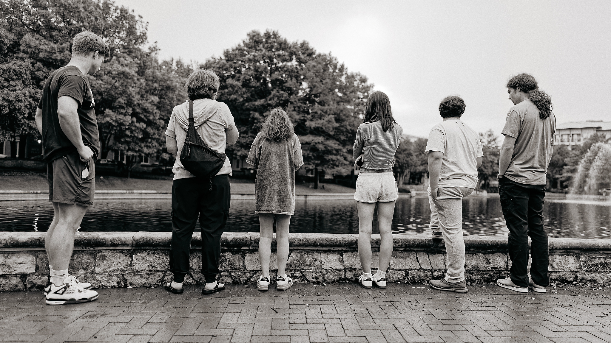 Six people stand in a row with their backs to the camera, looking over a stone wall at a pond with a fountain and trees.