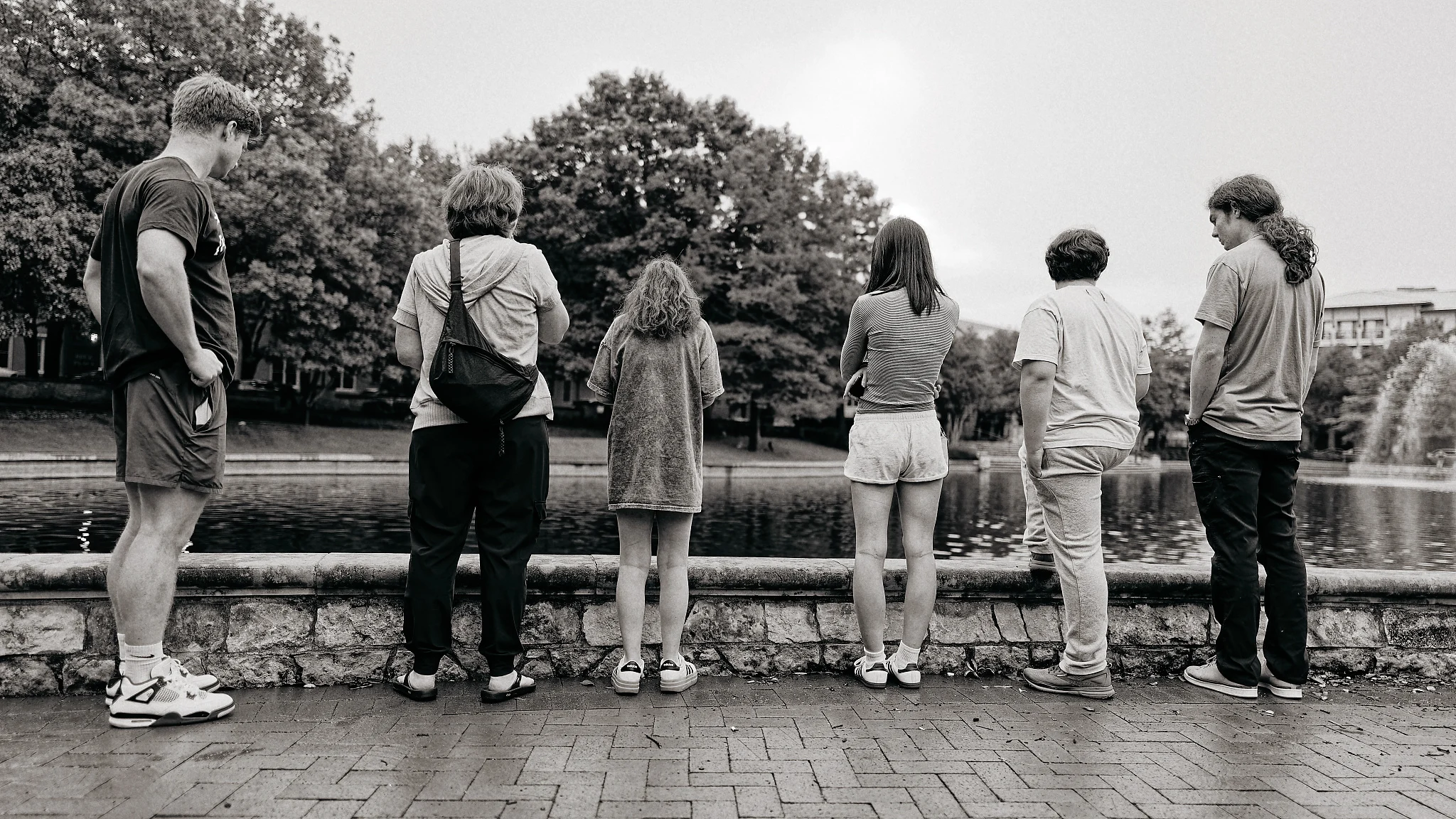 Six people stand in a row with their backs to the camera, looking over a stone wall at a pond with a fountain and trees.