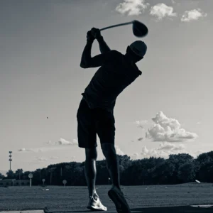 Man hitting a golf ball at the driving range silhouetted against a clear sky with a couple of clouds