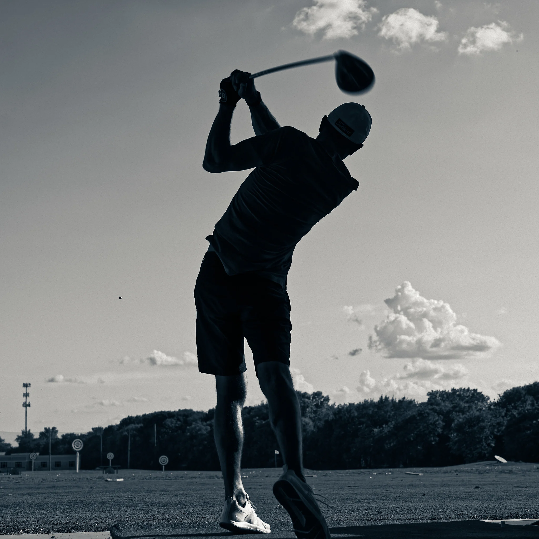 Man hitting a golf ball at the driving range silhouetted against a clear sky with a couple of clouds