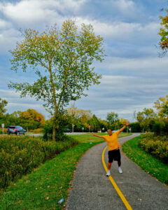 A smiling man in a yellow shirt and black shorts stands with arms outstretched on a paved path with a yellow center line, surrounded by trees and grass.