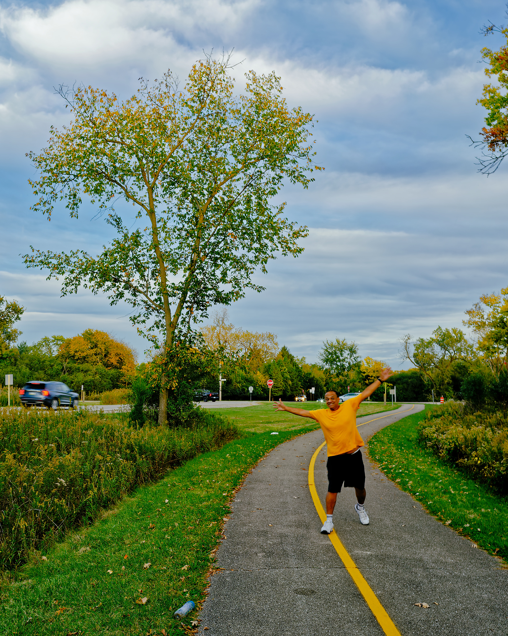 A smiling man in a yellow shirt and black shorts stands with arms outstretched on a paved path with a yellow center line, surrounded by trees and grass.
