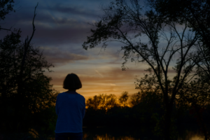 Woman standing with her back to the camera and arms crossed. She is looking out over a sunset with the shadows of the trees framing her silhouette