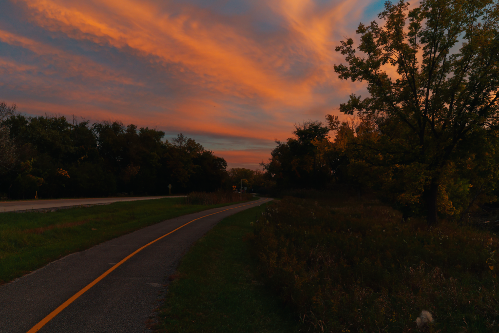 Bike path with a yellow lane stripe next to a road at the end of the day. Curving from the bottom to the right with trees alongside it and the sunset clouds curving to match the flow of the path.