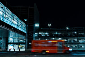 A blurred orange shuttle bus speeds past a glass-walled parking garage lit up at night.